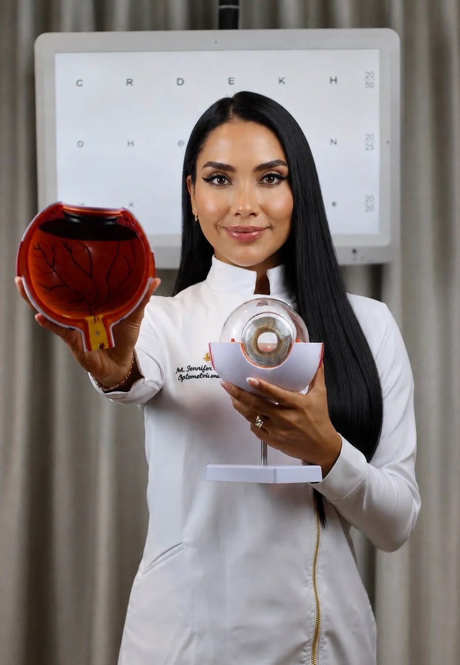Dra. Jennifer Moncayo sosteniendo un modelo anatómico del ojo y un lente frente a una carta optométrica, explicando el examen visual